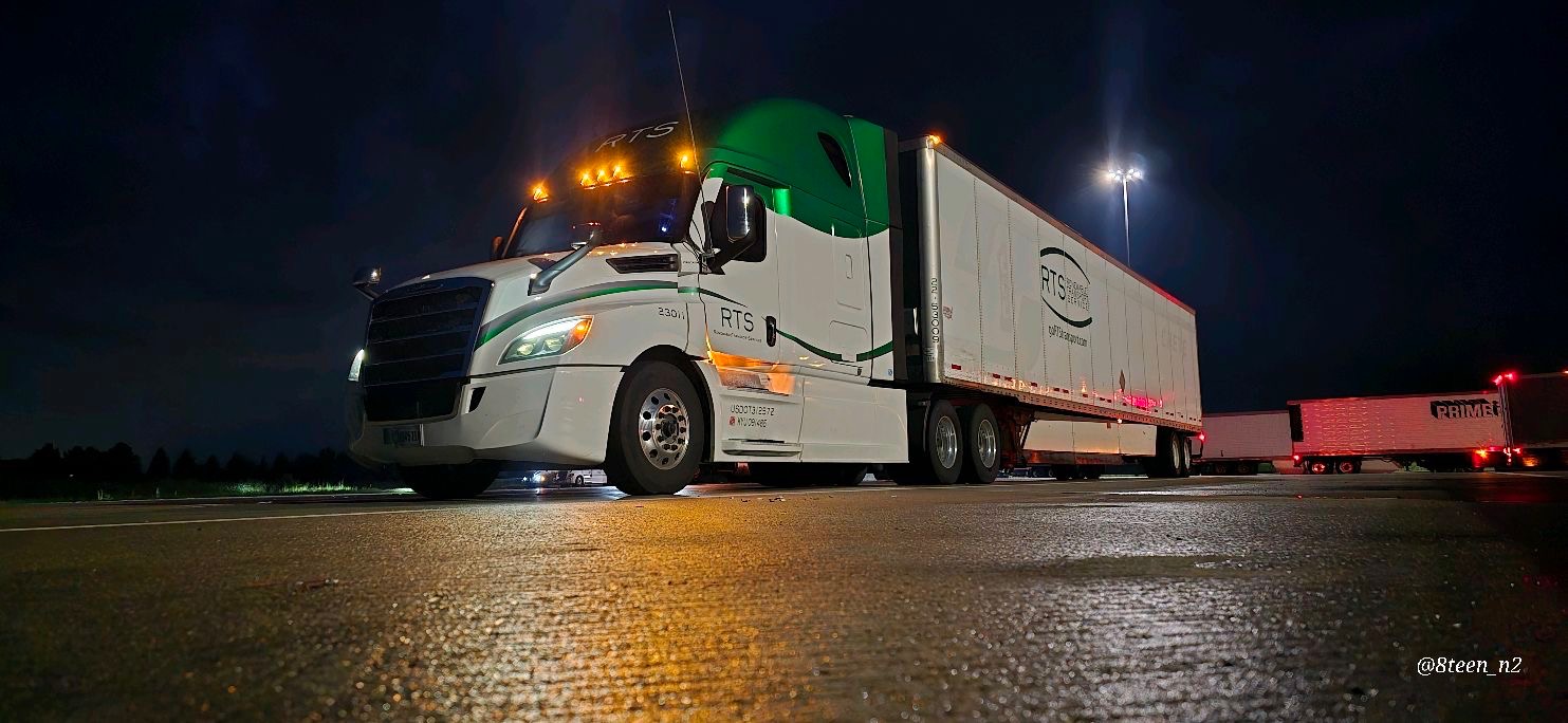 An RTS Freightliner Cascadia and reefer trailer lit up at a truck stop at night, reflecting on wet pavement